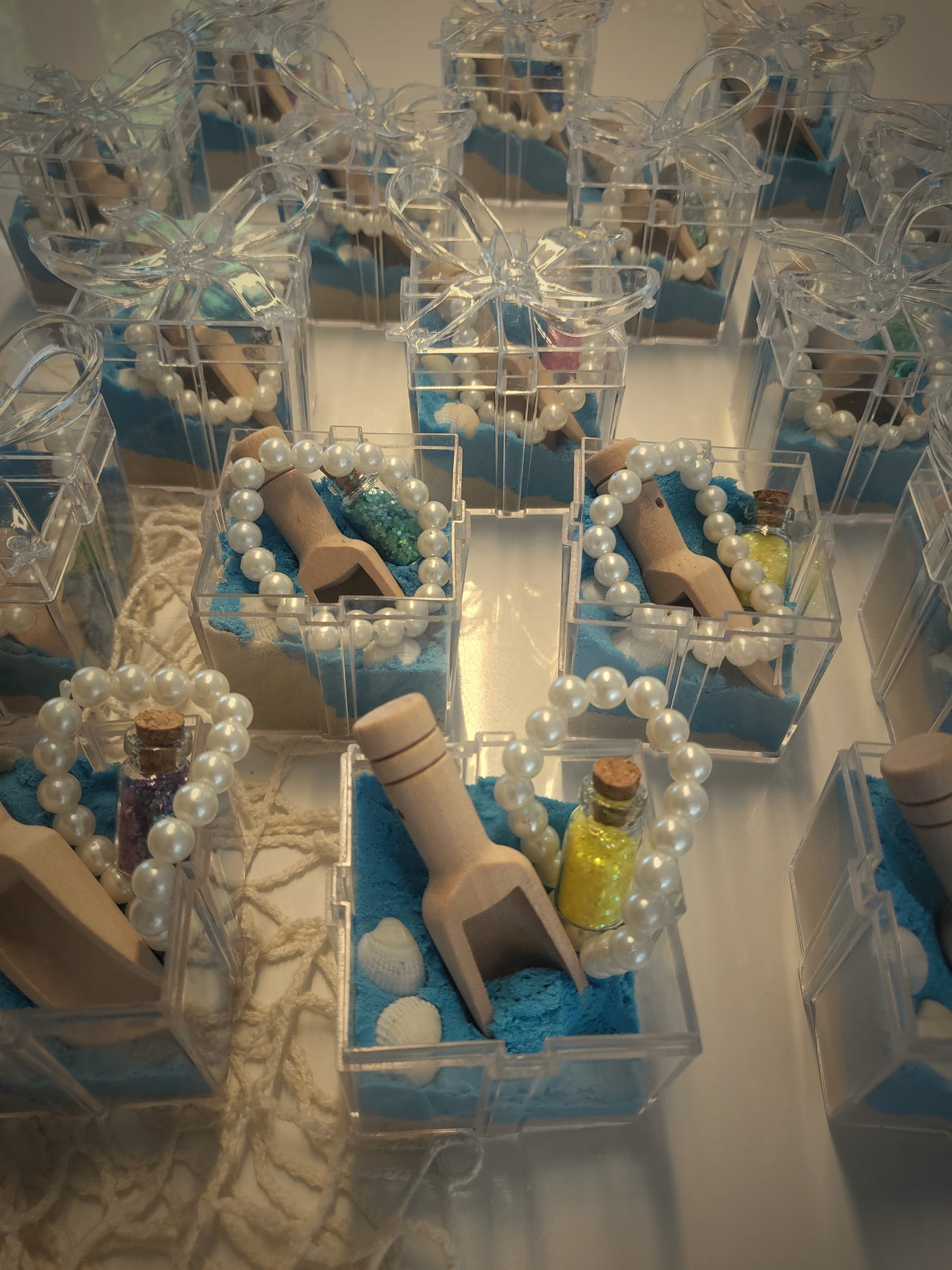 Lots of beautiful clear boxes filled with blue and natural kinetic sand and sea shells, pearl bracelets and a message in the bottle glitter. Sensory play Party favours